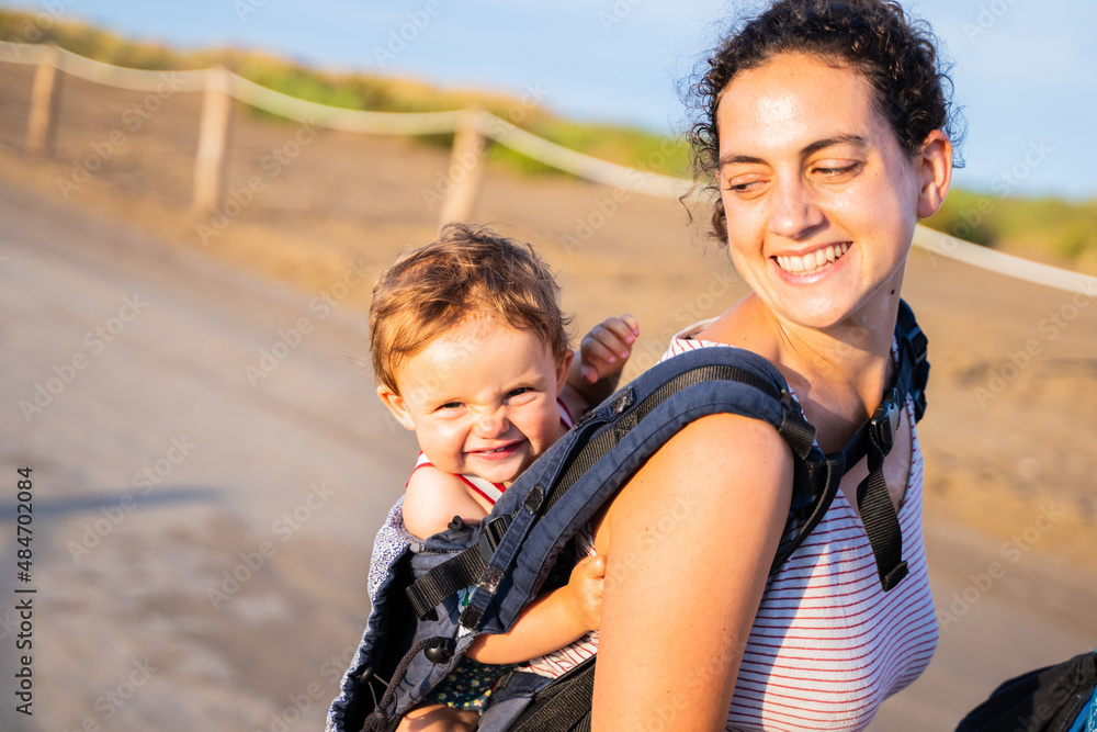 Happy mother carrying a smiling baby on her back in a baby carrier outdoors Happy mother carrying a smiling baby on her back in a baby carrier outdoors