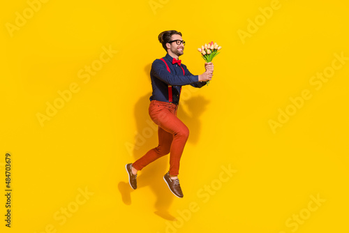 Full size profile side photo of young guy jump up hold bunch of tulips present look empty space isolated over yellow color background