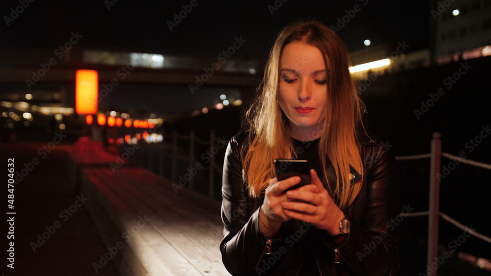 Young woman sitting on a red-lit street using a smartphone and smiling