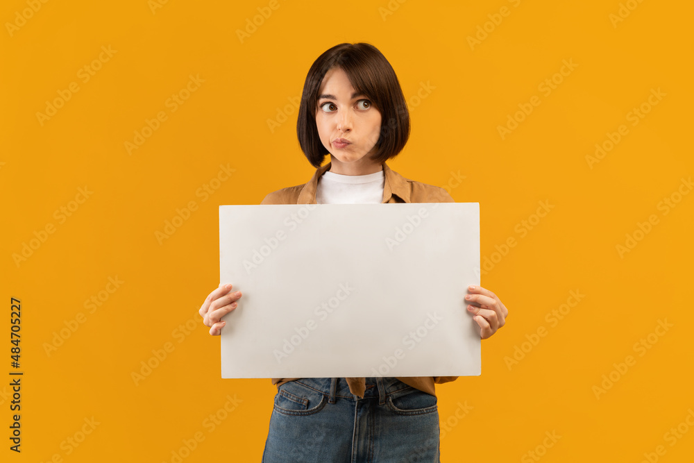 Making decision. Doubtful young lady holding empty blank board, posing on yellow studio background Making decision. Doubtful young lady holding empty blank board, posing on yellow studio background