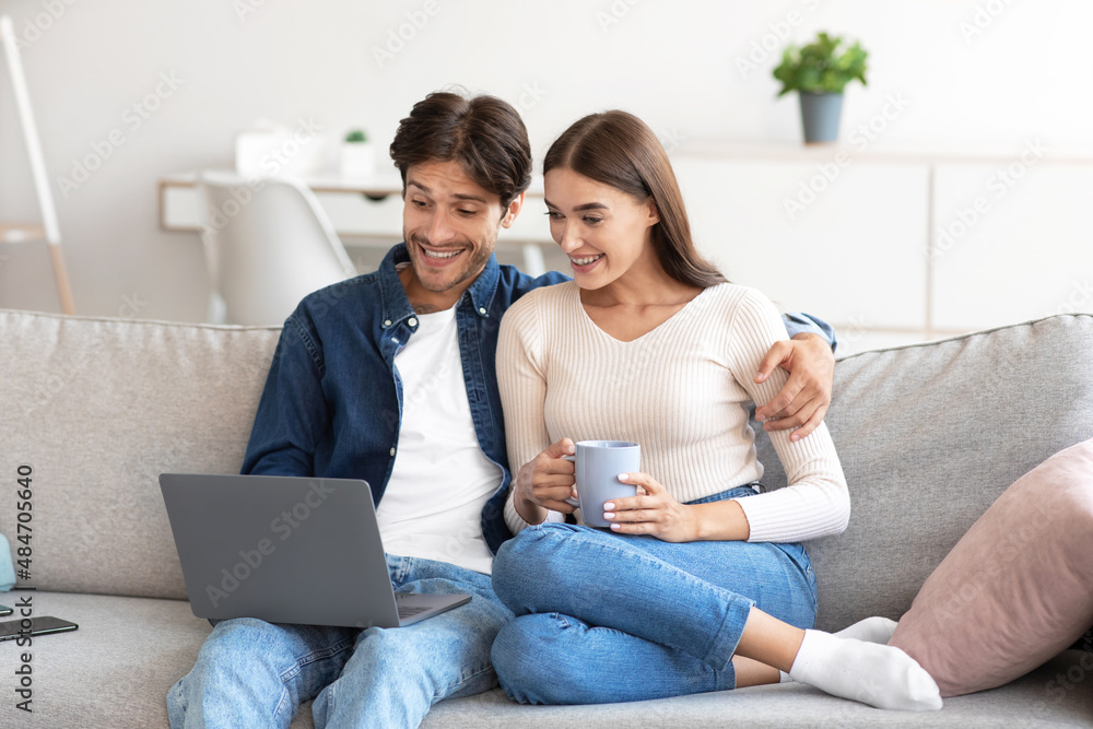 Shocking smiling millennial european couple sitting on couch looking at laptop, shopping, calling online Shocking smiling millennial european couple sitting on couch looking at laptop, shopping, calling online