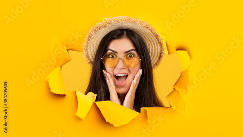 Happy surprised young latin female with open mouth in sunglasses and hat looks through hole in yellow paper © Prostock-studio