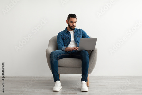 Full length of Arab man using laptop for online work, studies or communication, sitting in armchair, white studio wall