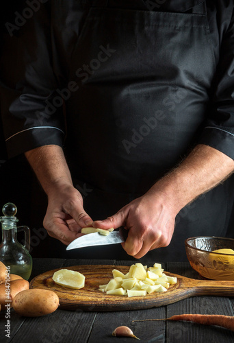 Chef cuts raw potatoes into pieces with a knife before preparing breakfast or dinner. Close-up of cook hands while working in restaurant kitchen