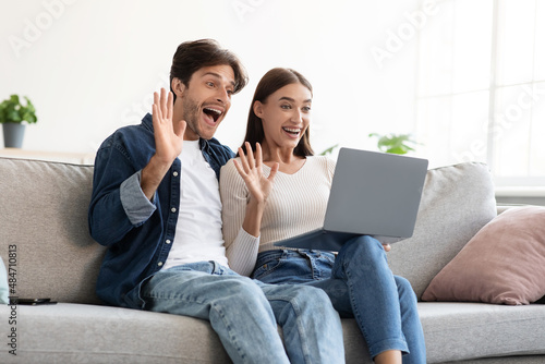 Satisfied young european guy and lady communicating with family remotely, waving hands to laptop webcam
