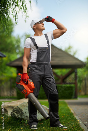 Εκτύπωση καμβά Full length portrait of handsome caucasian man in overalls, cap and gloves standing at garden with leaf blower in hands