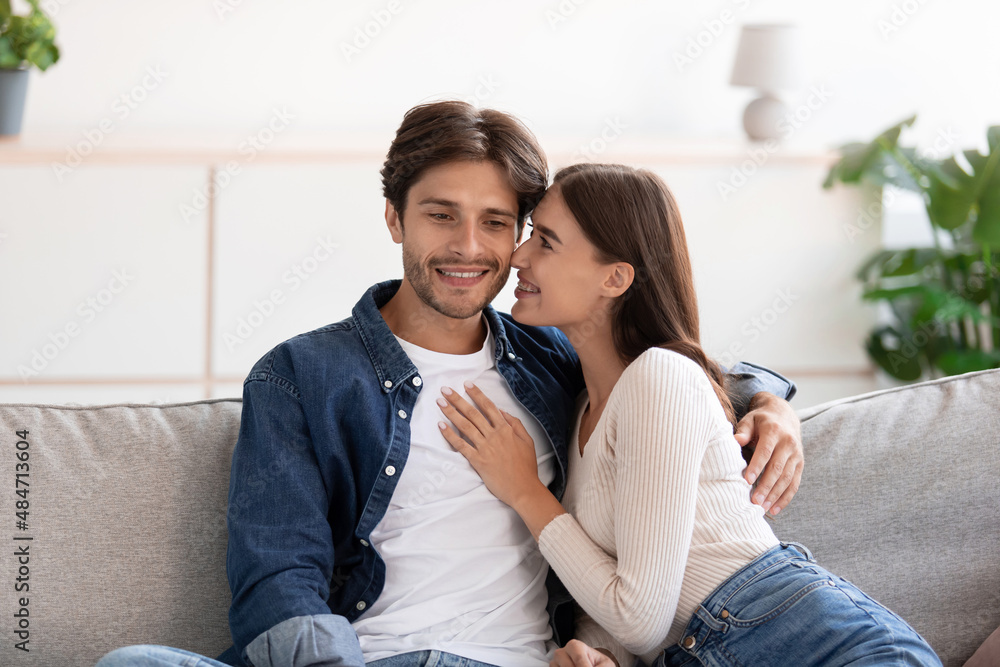 Fototapeta premium Cheerful millennial european woman kissing happy guy on cheek sitting on couch, look at camera