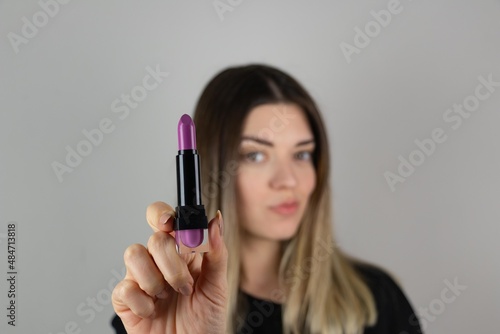 Close up photo of beautiful girl hold purple pomade. She will check new lips stick on isolated over gray background. Matte effect. Cosmetic, beauty and make up. Focus is on purple lipstick. 