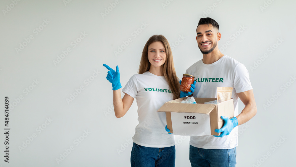 Millennial volunteers in protective masks and gloves with food donations box, woman pointing aside at copy space