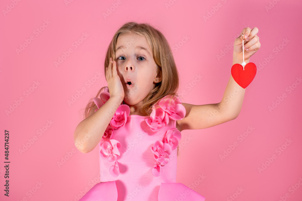 Portrait of surprised cute little blue-eyed girl with long fair hair in chic pink dress stand open-mouthed holding red paper heart on string on pink background. Valentines day, love, emotions concept. Portrait of surprised cute little blue-eyed girl with long fair hair in chic pink dress stand open-mouthed holding red paper heart on string on pink background. Valentines day, love, emotions concept.