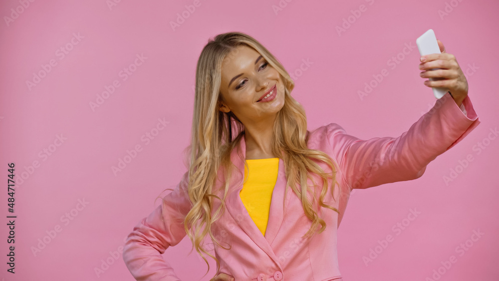 Positive blonde woman in jacket taking selfie on cellphone isolated on pink. Positive blonde woman in jacket taking selfie on cellphone isolated on pink.
