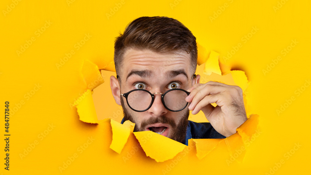 Surprised millennial caucasian guy takes off his glasses and looks in camera through hole in yellow paper Surprised millennial caucasian guy takes off his glasses and looks in camera through hole in yellow paper