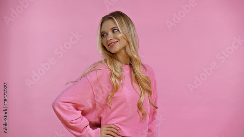 Smiling woman in sweatshirt holding hand on hip isolated on pink.