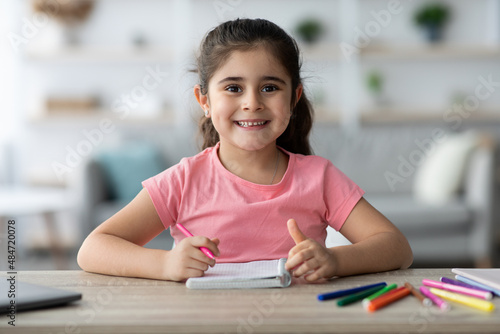 Cute Little Arab Girl Sitting At Desk At Home, Drawing In Notepad