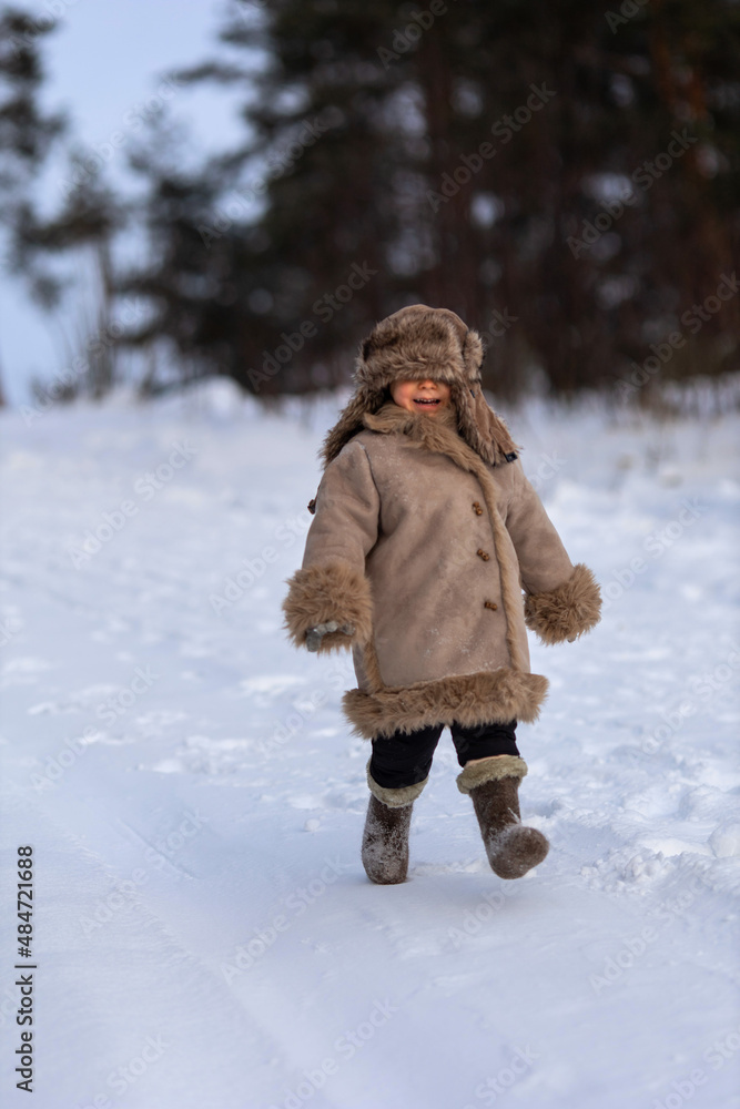 A kid in a winter fur coat, hat and felt boots in a snow-covered forest A kid in a winter fur coat, hat and felt boots in a snow-covered forest