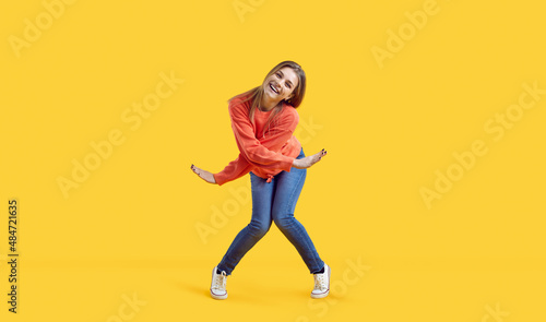 Funny carefree beautiful young woman having fun. Happy pretty teenage girl in comfortable casual wear looking at camera and smiling while dancing in the studio against vibrant yellow colour background