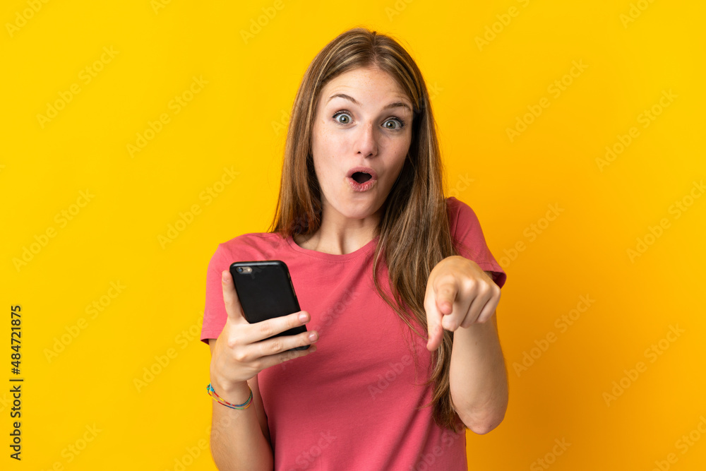 Young woman using mobile phone isolated on yellow background surprised and pointing front Young woman using mobile phone isolated on yellow background surprised and pointing front