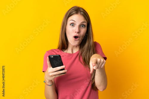 Young woman using mobile phone isolated on yellow background surprised and pointing front