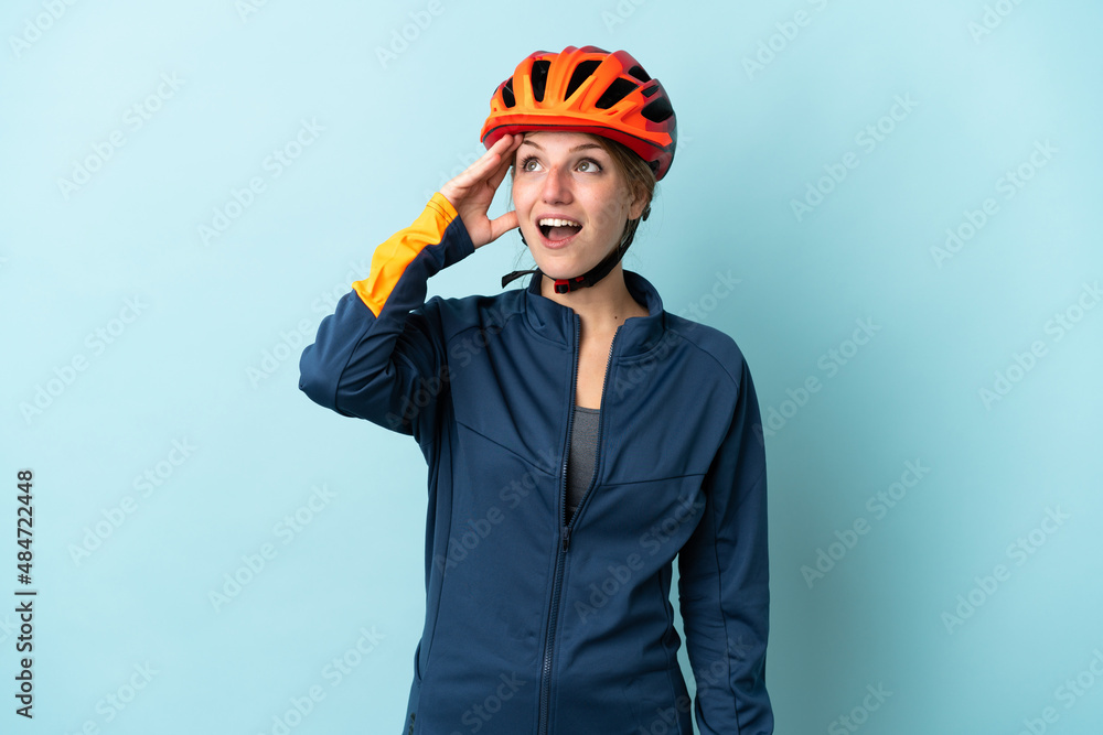Young cyclist woman isolated on blue background doing surprise gesture while looking to the side - obrazy, fototapety, plakaty 