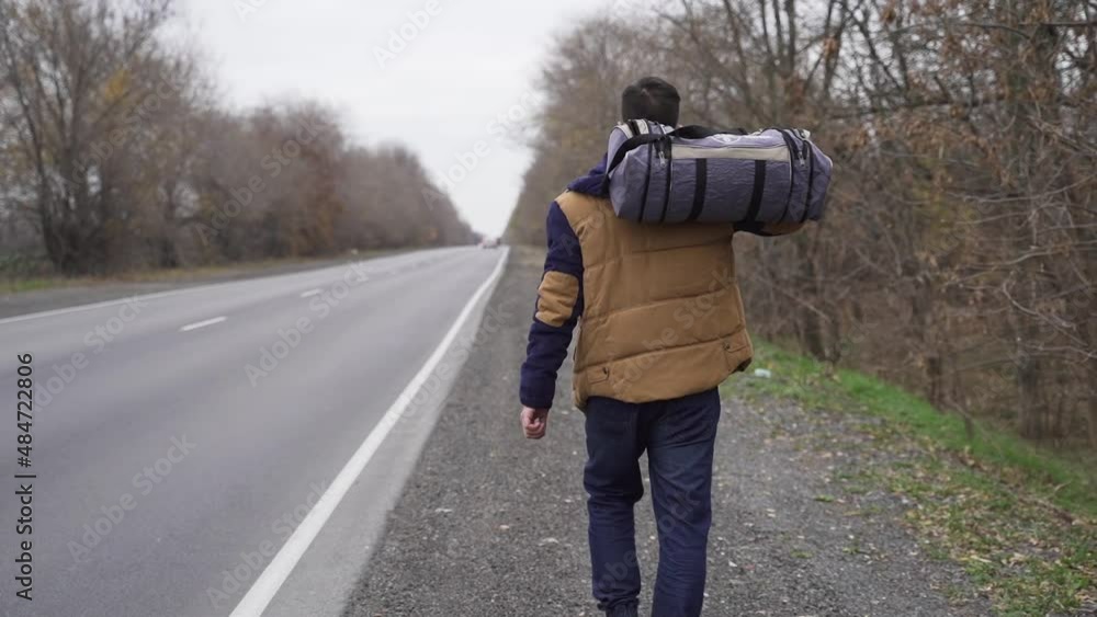 Guy with bag is walking along the track with bag. Traveler with things walks along the roadside during the day Guy with bag is walking along the track with bag. Traveler with things walks along the roadside during the day