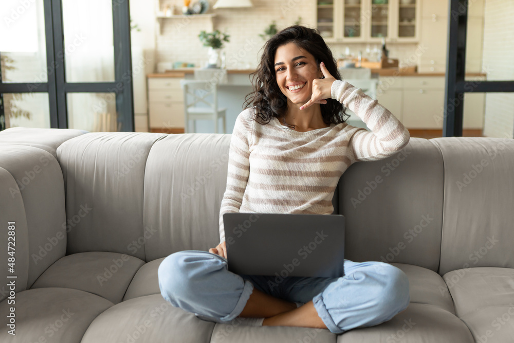 Cheerful young woman doing phone gesture, saying call me back, using laptop, sitting on couch at home, copy space Cheerful young woman doing phone gesture, saying call me back, using laptop, sitting on couch at home, copy space