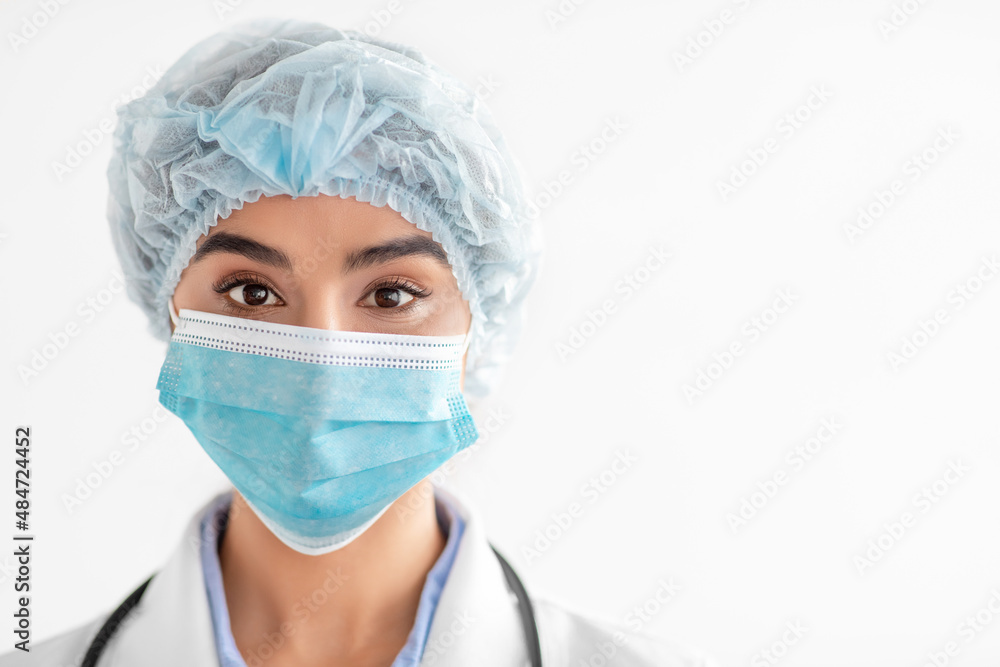 Portrait of millennial indian lady doctor in coat, protective mask and cap, looking at camera Portrait of millennial indian lady doctor in coat, protective mask and cap, looking at camera