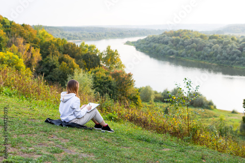 A student girl is sitting on the river bank and writing in a notebook