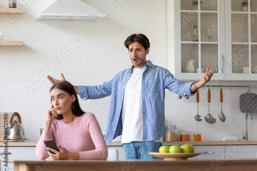 Sad young european woman with smartphone ignores offended angry screaming gesticulating husband