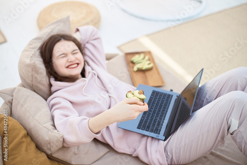 Young smiling woman is eating toast with avocado and working on laptop