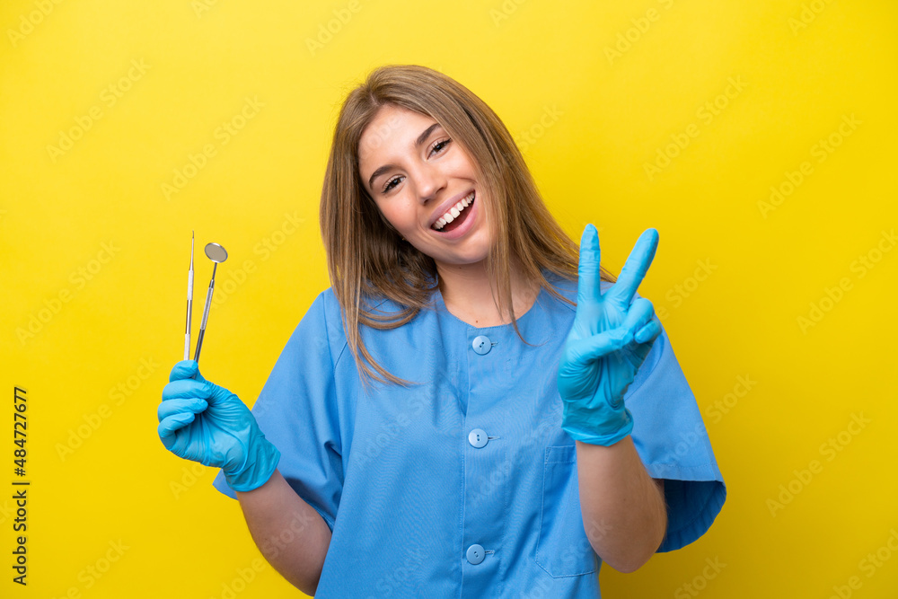 Dentist caucasian woman holding tools isolated on yellow background smiling and showing victory sign Dentist caucasian woman holding tools isolated on yellow background smiling and showing victory sign