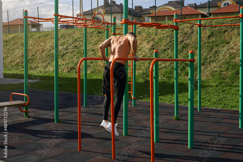 Back view of shirtless caucasian man exercising on parallel bars outdoors. Strong male athlete doing doing triceps dip on sports ground. Bodybuilding concept.