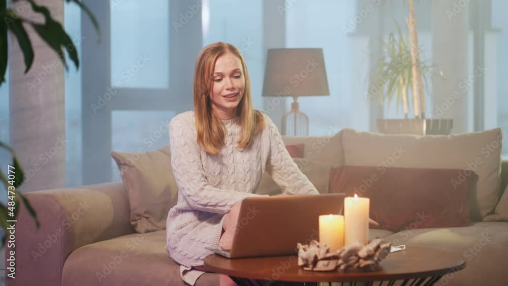Beautiful young woman with blond hair having video call on wireless laptop while sitting on couch. Dark atmosphere at modern apartment and burning candles on table. Beautiful young woman with blond hair having video call on wireless laptop while sitting on couch. Dark atmosphere at modern apartment and burning candles on table.