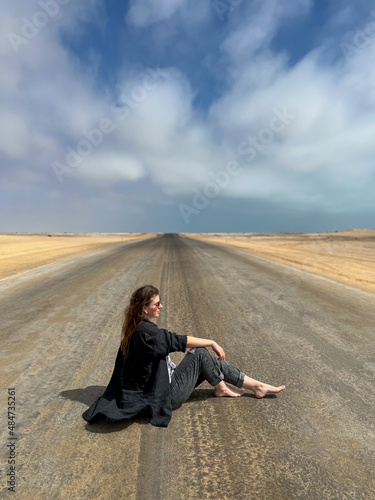 Young woman sits on highway in desert. Girl on center of the road, barefoot.