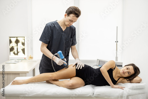 Doctor applies shock wave therapy with special medical equipment on women's thigh muscle at medical office. Concept of non-invasive technology for treating pain in musculoskeletal system
