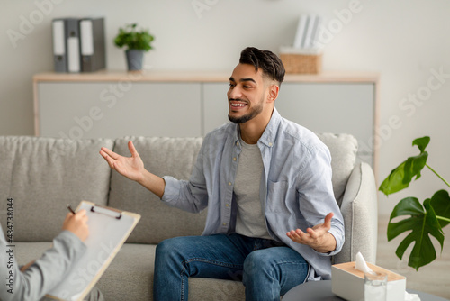 Young happy arab guy talking to his psychologist, receiving professional help, sitting on sofa at mental health clinic photo