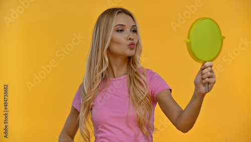Blonde woman pouting lips while holding mirror isolated on yellow. © LIGHTFIELD STUDIOS
