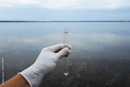  The scientist is taking water sample of sea water. Test tube with water in a hand. Sea water pollution concept. Cloudy sky.....