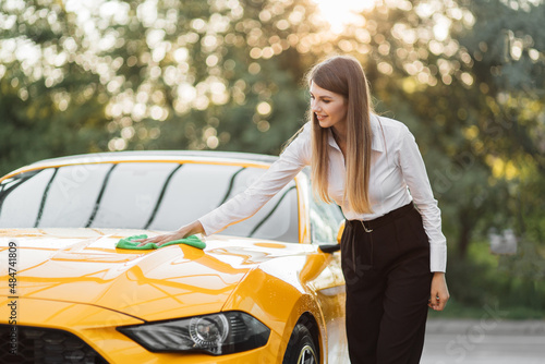 Portrait of a smiling young caucasian businesswoman on white shirt and black pant, cleaning modern sport car hood with green microfiber cloth outdoors, at the self car wash station. © sofiko14
