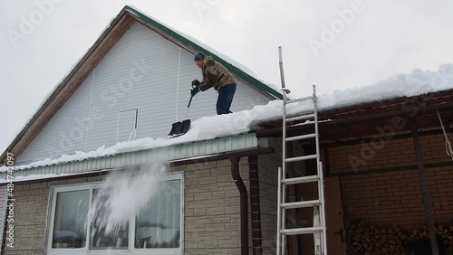 a man removes snow from the roof of a completely snow-covered house with a shovel. a lot of fresh snow after a blizzard, hard and dangerous work
