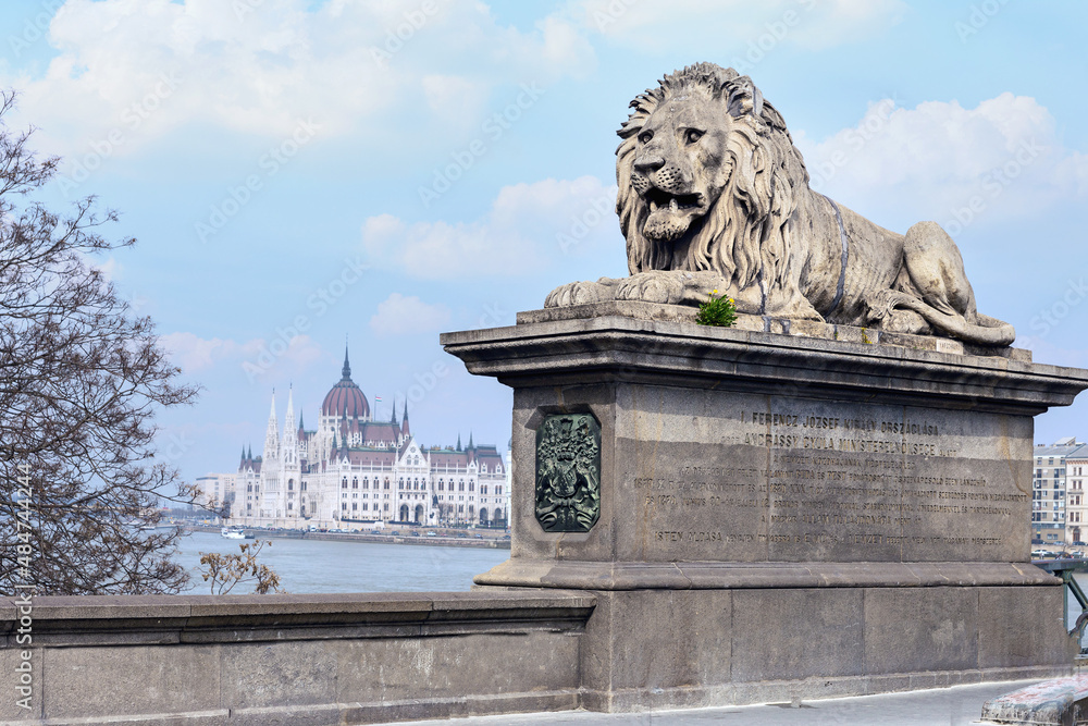 a concrete sculpture of a formidable lion on the railing of a bridge over the Danube River with the building of the Hungarian Parliament in the background