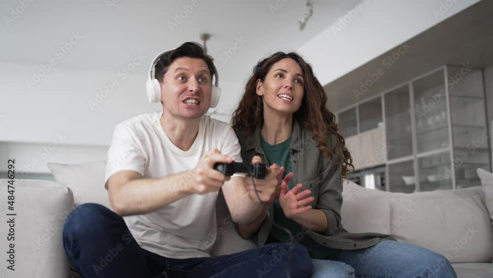 Newlyweds of worried brunette woman and excited man in wireless headphones sit on sofa happy from win in play station game Newlyweds of worried brunette woman and excited man in wireless headphones sit on sofa happy from win in play station game