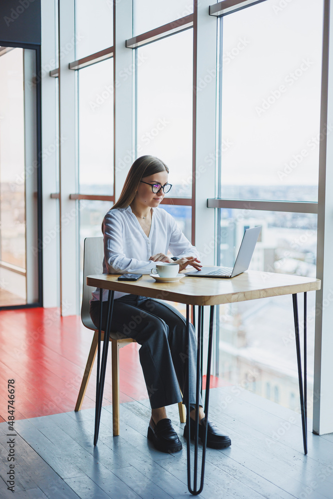 Happy young woman manager in casual attire and glasses smiles and looks into a laptop, looking through different information, she drinks coffee in a cozy cafe with large windows and a view of the city Happy young woman manager in casual attire and glasses smiles and looks into a laptop, looking through different information, she drinks coffee in a cozy cafe with large windows and a view of the city