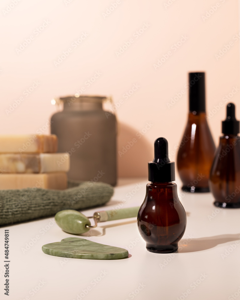 Amber glass spray and dropper bottles with cosmetic on light bathroom table. Natural jade roller for facial massage and Guasha Massage Board. Amber glass spray and dropper bottles with cosmetic on light bathroom table. Natural jade roller for facial massage and Guasha Massage Board.