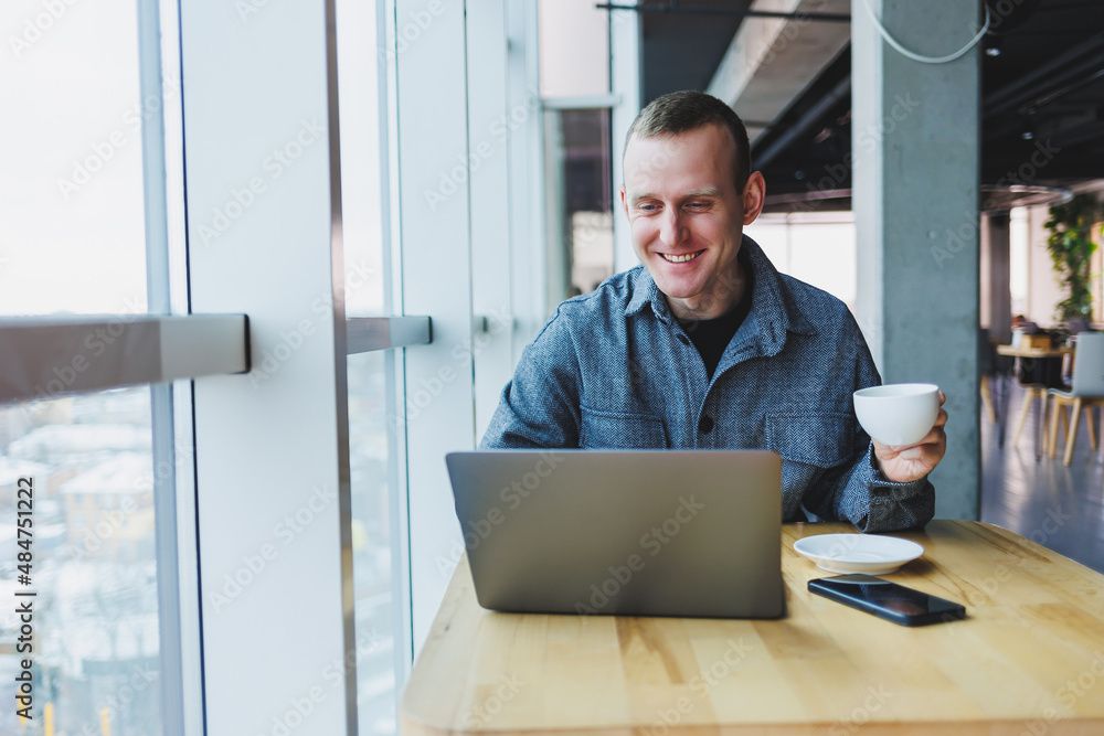 Successful happy business man is sitting at a table in a cafe, holding a cup of coffee and using a laptop. Successful happy business man is sitting at a table in a cafe, holding a cup of coffee and using a laptop.