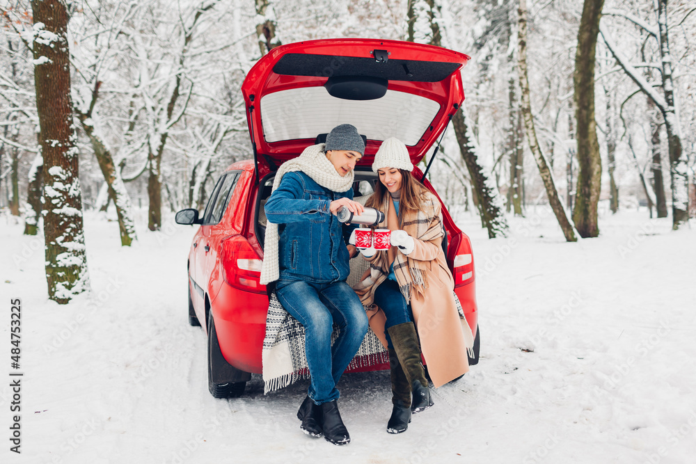 Valentine's day couple in love sitting in car trunk drinking hot tea in snowy winter forest. People relaxing during date
