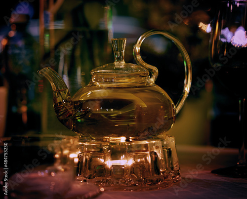 Transparent teapot with green tea on a heated glass stand on a restaurant table with rays of reflective illumination
