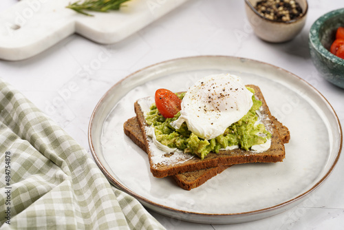 Trendy lifestyle sandwich: protein bread slice with cream cheese, mashed avocado, cherry tomatoes and poached egg on white scandi plate, light setting