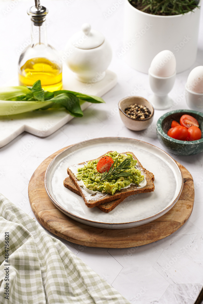 Trendy lifestyle sandwich: protein bread slice with cream cheese, mashed avocado, cherry tomatoes and rosemary on white scandi plate, light setting