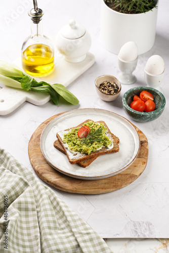 Trendy lifestyle sandwich: protein bread slice with cream cheese, mashed avocado, cherry tomatoes and rosemary on white scandi plate, light setting