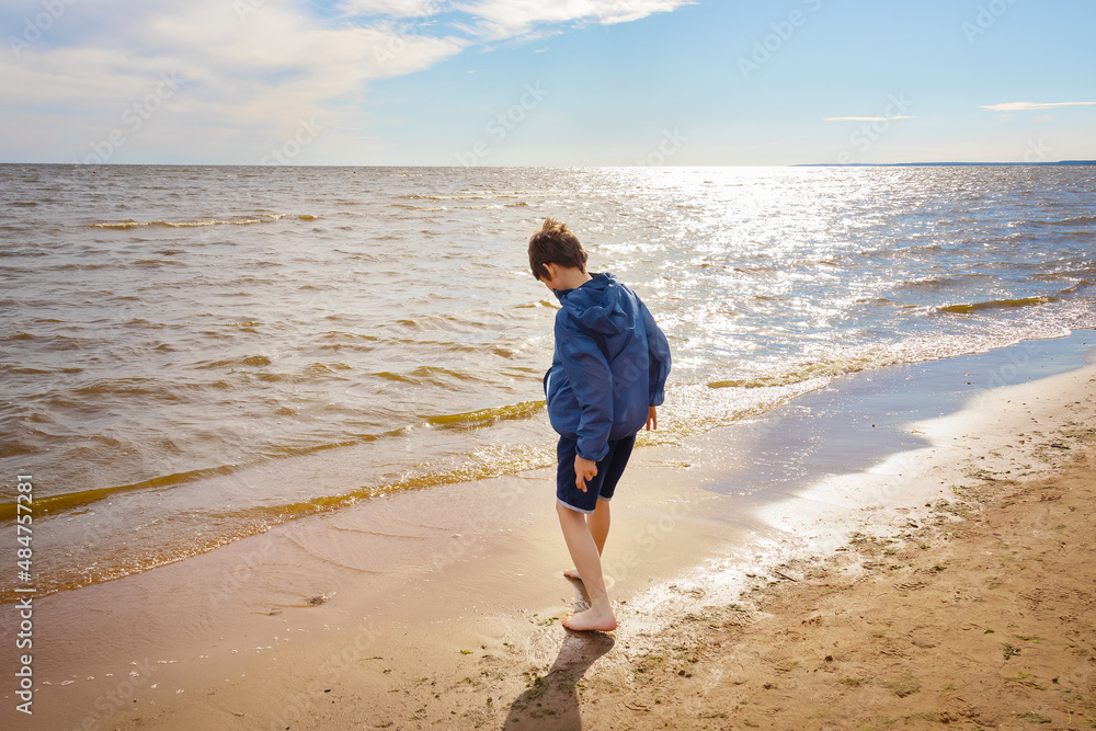 Barefoot boy on surf line on sunny beach standing with his back in blue jacket and shorts